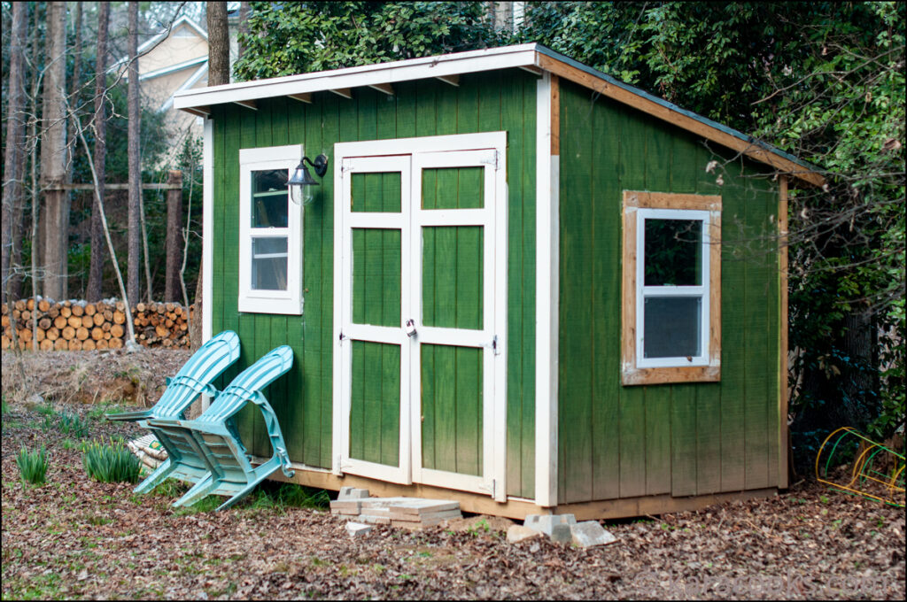 A green garden shed in a wooded area with a couple of blue chairs leaned up against the front